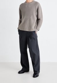 Helles braun gerippter Pullover mit einem diagonalen Muster, kombiniert mit locker sitzenden schwarzen Hosen und schwarzen Lederschuhen.