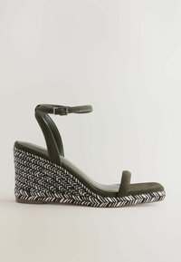 Vihreät mokkanahkakengät, joissa on valko-navy herringbone-kuvioinen korko. Varustettu säädettävällä nilkkahihnalla ja avonaisella varpaalla.