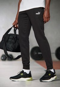 Black athletic pants with white Puma logo, paired with black sneakers featuring green accents, and a black gym bag with a mesh pocket.