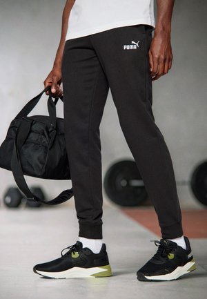 Schwarze Sporthose mit weißem Puma-Logo, kombiniert mit schwarzen Sneakers mit grünen Akzenten und einer schwarzen Sporttasche mit einem Netzfach.