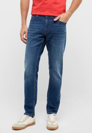 Man wearing blue slim-fit jeans, red shirt, and white sneakers standing with one hand in pocket against plain background.