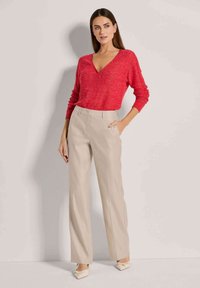 Rode v-hals trui, lange mouwen, gedragen met beige, wijde broekspijpen. Lichte stofstructuur, platte schoenen met linten aan de achterkant.