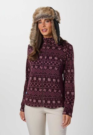 Une femme porte une veste polaire bordeaux à motifs et à fermeture éclair, un pantalon beige et un chapeau trappeur marron doublé de fourrure, sur un fond uni.