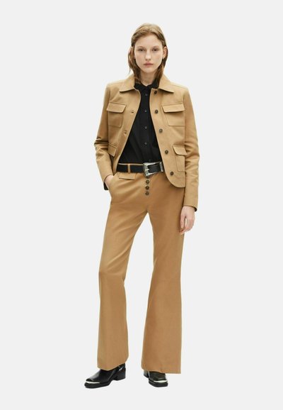 Veste en daim tan avec poches à rabat, associée à un pantalon beige sur mesure et une chemise noire. Des bottes noires à talons et une ceinture en argent rehaussent la tenue.
