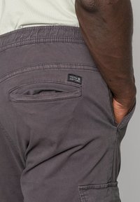 Grijze cargoshorts met een trekkoord in de taille, voorzien van een zijzak en een label met de tekst "NINETEEN NINETY THREE" nabij de achterzak.