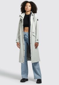 Light gray waterproof coat with a hood, snap button closure, side pockets, and a mesh lining, paired with loose blue jeans and black shoes.