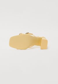 Yellow heeled sandal with a wide block heel, textured sole, and thin, crisscross straps. Features a buckle accent on the ankle strap.