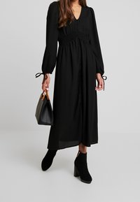 Schwarzes langärmliges Kleid mit V-Ausschnitt, gerafftem Taillenbereich und Knopfleiste. Kombiniert mit schwarzen Stiefeletten und einer kleinen schwarzen Handtasche.