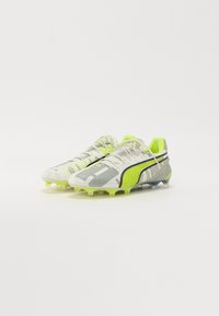 Botas de fútbol en blanco, gris y amarillo neón. Presentan una parte superior texturizada, patrones intrincados y una suela lisa con múltiples clavos para tracción.