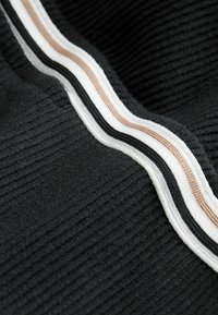 Close-up van zwart geribd stof met drie verhoogde strepen in wit, zwart en beige, die diagonaal over het oppervlak lopen.