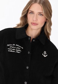 Czarna kurtka z korduroju z zapięciem na srebrne guziki, haftowanym napisem po lewej stronie oraz wzorem kotwicy na prawej piersi.