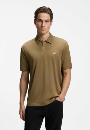 Junger Mann mit kurzen braunen Haaren, der ein braunes Porsche BOSS Poloshirt und schwarze Hose trägt, steht mit einer Hand in der Tasche.