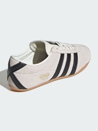 Suede tenisice u svijetlosivoj boji s crnim prugama i detaljima. Imaju smeđu gumenu podlogu i zlatni logo s strane.