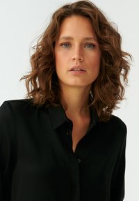 Camicia nera con colletto, bottoni frontali, maniche corte e texture liscia. La pelle circostante ha tonalità chiare e un aspetto naturale.