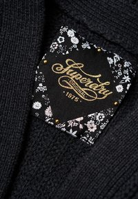 Černá pletenina s texturovaným vzorem; má štítek s bílým květinovým designem a zlatým textem "Superdry Original 1975".