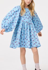 Lys blå kjole med puffede ærmer, med et gentaget print af enhjørninger i lilla og hvid. Let flydende design med samlet talje.