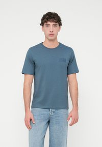 Μπλουζάκι σκούρου μπλε με κοντά μανίκια από απαλό ύφασμα, με στρογγυλή λαιμόκοψη. Διαθέτει διακριτικό λογότυπο τυπωμένο στο αριστερό στήθος. Φορώντας το με ανοιχτόχρωμο μπλε τζιν.