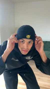 Czarna czapka beanie z żółtym graficznym logo, noszona przez osobę, która ją poprawia. W tle widać jasną ścianę oraz zieloną kanapę.