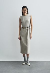 Μπεζ αμάνικο φόρεμα με στενή γραμμή, σφιγμένο στη μέση με μαύρη ζώνη. Το μοντέλο φορά γκρι κάλτσες και μυτερές λευκές γόβες.