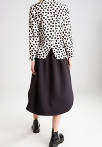 Schwarz-weiße Bluse mit Herzmuster und langen Ärmeln, mit einem Schlitz hinten, kombiniert mit einem schwarzen Midirock und glänzenden schwarzen Stiefeletten.