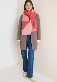 Roze en rode sjaal met geometrisch patroon en textuurbreiwerk, gedragen over een grijze jas, gecombineerd met een blauwe spijkerbroek en witte sneakers.