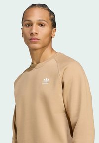 Sudadera beige hecha de tela suave, con un cuello redondo y mangas raglán. Logo blanco de Adidas en el lado izquierdo del pecho. Textura suave.