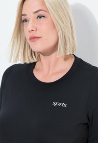 Zwart sportshirt met een ronde hals en korte mouwen, met een wit 'sport' logo op de bovenste linker borst.