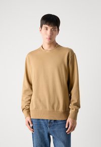 Sudadera marrón hecha de una tela suave, con cuello redondo, puños y dobladillo acanalados. Llevada con jeans de mezclilla azules.