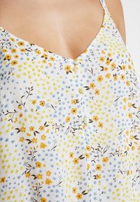 Blusa con stampa floreale caratterizzata da uno sfondo bianco con fiori gialli, blu e marroni. Ha una scollatura a V e un dettaglio centrale con bottoni.