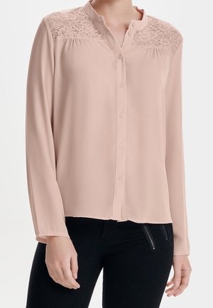 Hellrosa Langarmbluse mit Spitzen-Details auf den Schultern, vorne mit Knöpfen, lockere Passform, getragen zu schwarzen Hosen mit Reißverschlusstaschen.