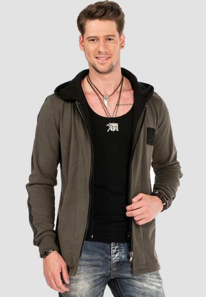 Olivgrüne Zip-Hoodie mit schwarzer Kapuze, taillierter Passform und gerippten Bündchen. Über einem schwarzen Tanktop mit silbernen Schmuckakzenten getragen.
