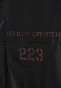 Sort jakke med teksten "OFF DUTY OFF PITCH" og nummeret "223" i mørk maroon. Glat tekstur med et minimalistisk design.