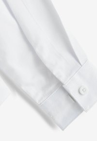 Manche de chemise en coton blanc avec fermeture par bouton, texture lisse et couture soignée. Le poignet est légèrement arrondi avec une finition nette.