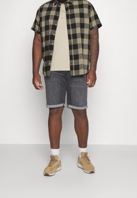 Camisa de cuadros de manga corta en oliva y negro, camiseta beige, pantalones cortos de mezclilla gris con dobladillo enrollado y zapatos deportivos marrones claros con calcetines blancos.