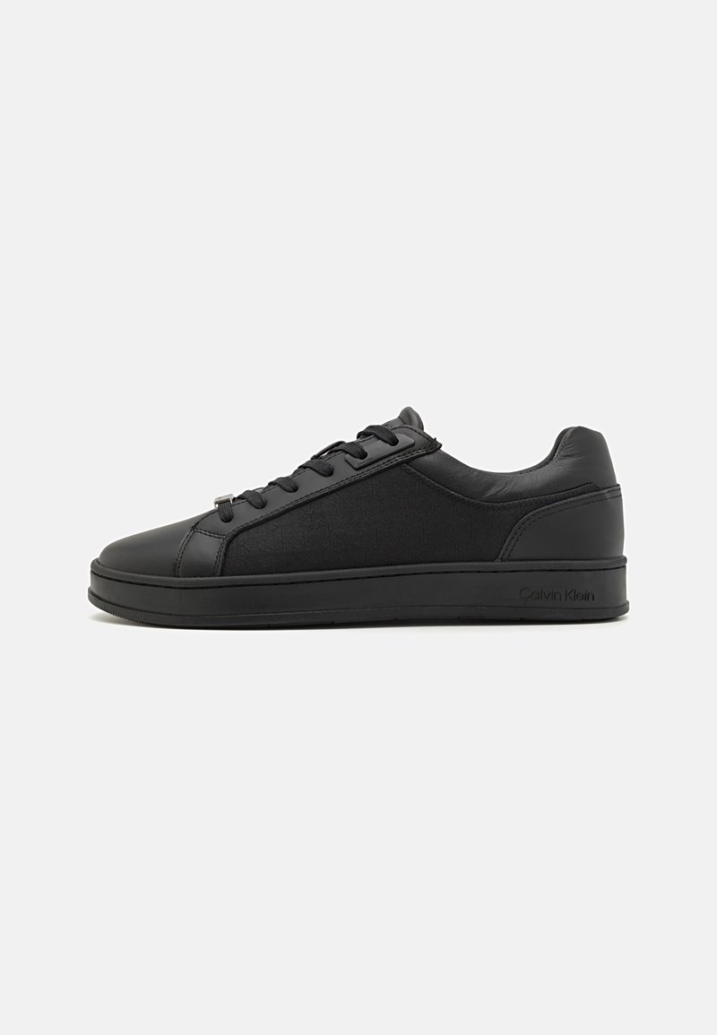 Black sneaker with a smooth leather upper, featuring a textured panel, round toe, and flat rubber sole. Laces and subtle logo detail present.