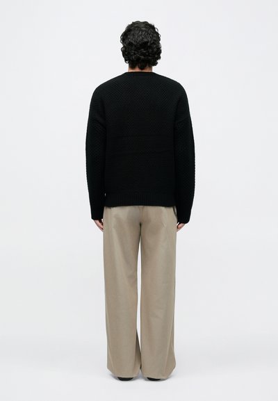 Άνδρας με σγουρά μαλλιά που φοράει μαύρο πουλόβερ με υφή και μπεζ παντελόνι φαρδύ, στέκεται με την πλάτη προς τα εμπρός σε απλό λευκό φόντο.
