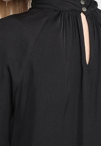 Blouse noire à col haut avec une ouverture en forme de clé fermée par deux boutons métalliques au niveau du col, mettant en valeur des détails de tissu froncé.