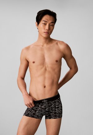 Jonge man met kort zwart haar die zwarte Calvin Klein boxershorts draagt met witte tekst op een effen grijze achtergrond.
