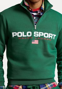 Personne portant un sweat-shirt vert à fermeture éclair quart avec « POLO SPORT RALPH LAUREN » et un logo du drapeau américain sur une chemise à carreaux multicolores.
