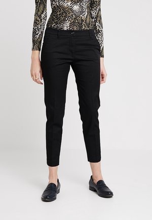 Kobieta ubrana w dopasowane czarne spodnie za kostkę, czarne mokasyny oraz długi rękaw z nadrukiem w węża, stojąca na tle jasnego, jednolitego tła.