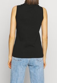 Kvinna som bär en ärmlös svart topp och ljusblå denimjeans, stående med ryggen vänd mot kameran framför en enkel bakgrund.