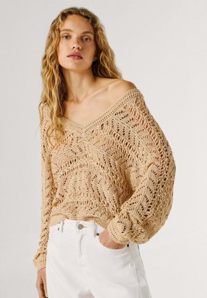 Mujer rubia con suéter beige de punto abierto y escote barco con mangas globo y pantalones blancos de talle alto, con una mano en el bolsillo.