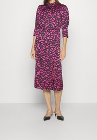 Zwart lange mouw jurk van stof met een roze lippenstift kusprint, met een hoge halslijn en een split aan de voorkant, gecombineerd met zwarte puntige schoenen.