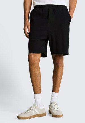 Man draagt zwarte geribbelde korte broek, witte sokken, beige sneakers, staat met handen in de zakken tegen een effen achtergrond.