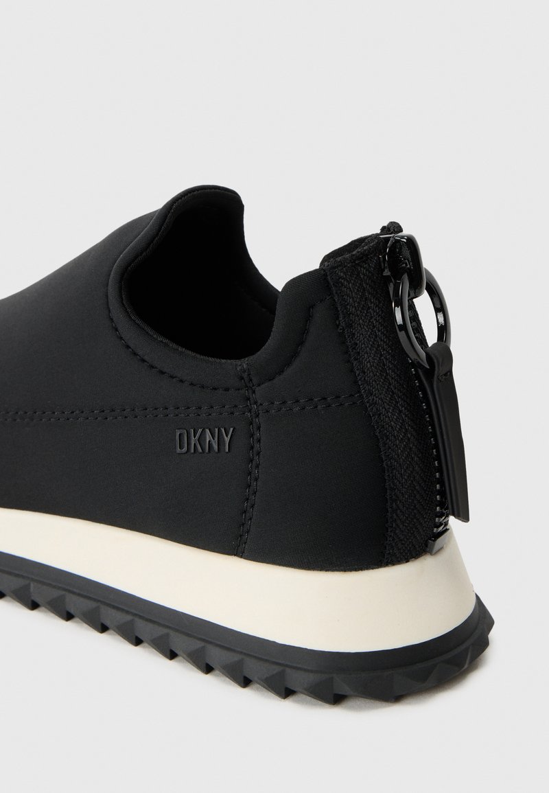 Chaussure noire à enfiler avec un tissu texturé, fermeture éclair sur le côté et une semelle en caoutchouc blanche contrastante avec un motif de bande de roulement en zigzag.