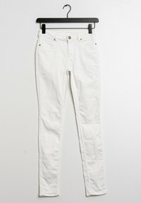 Jeans skinny de mezclilla blanca en un perchero negro. Cuenta con un cierre de botón frontal, cinco bolsillos y una tela suave, ligeramente texturizada.