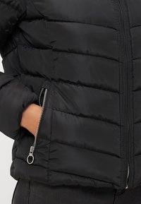 Casaco puffer preto com seções acolchoadas, um bolso lateral com zipper e uma textura suave, projetado para calor e conforto.