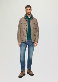 Beige jas met vier voorzakken, groene hoodie eronder, donkerblauwe jeans en bruine laarzen. Glad textuur en casual ontwerp.