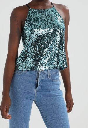 Migoczący turkusowy top na szyję z cekinów, zestawiony z wysokim stanem niebieskich dżinsów na stojącej osobie na jednolitym tle.