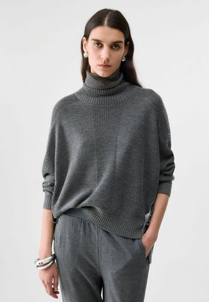 Femme aux longs cheveux foncés porte un pull en maille gris à col montant et un pantalon gris assorti, accessoirisés de larges boucles d’oreilles et bracelets argentés.